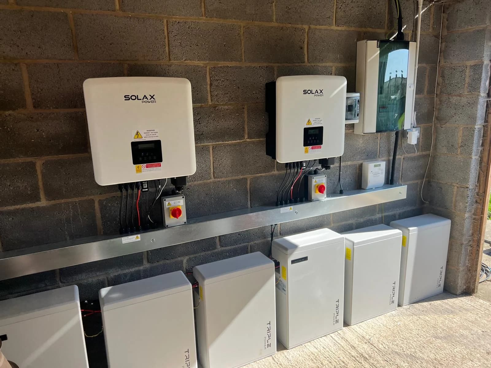 Two SolaX inverters with red isolators above six Triple Power battery modules on a bare blockwork wall