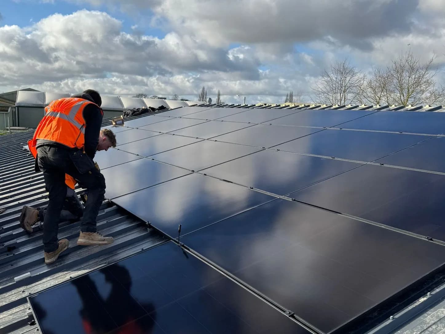 Two installers in orange high visibility vests fixing all black solar panels to a metal commercial roof