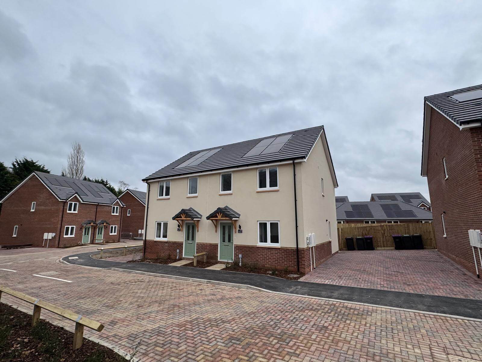 A cream rendered pair of new semi detached homes with in roof solar panels, beside a block paved close