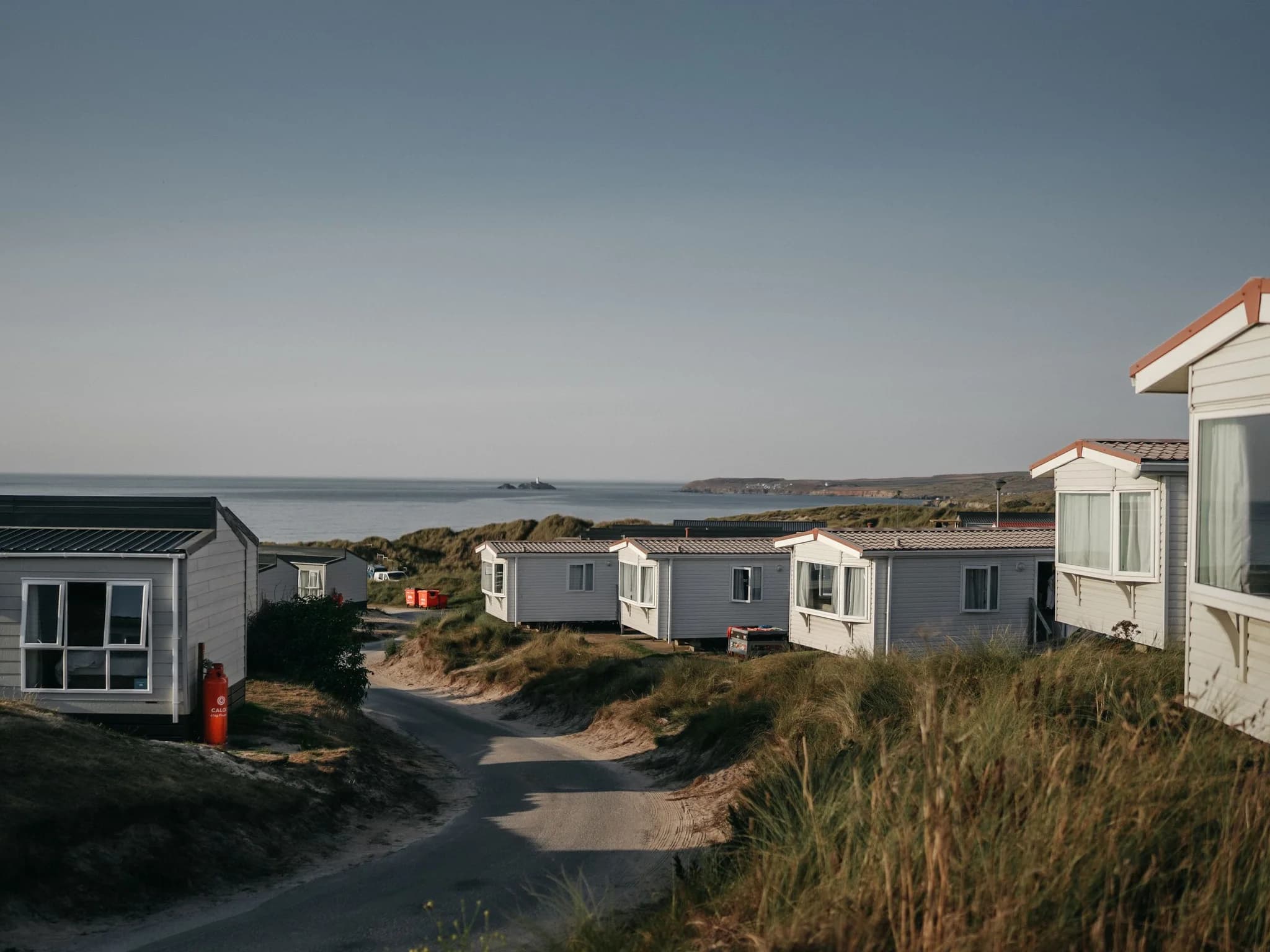 Static caravans along a sandy lane through dune grass, with the sea and a small lighthouse island beyond