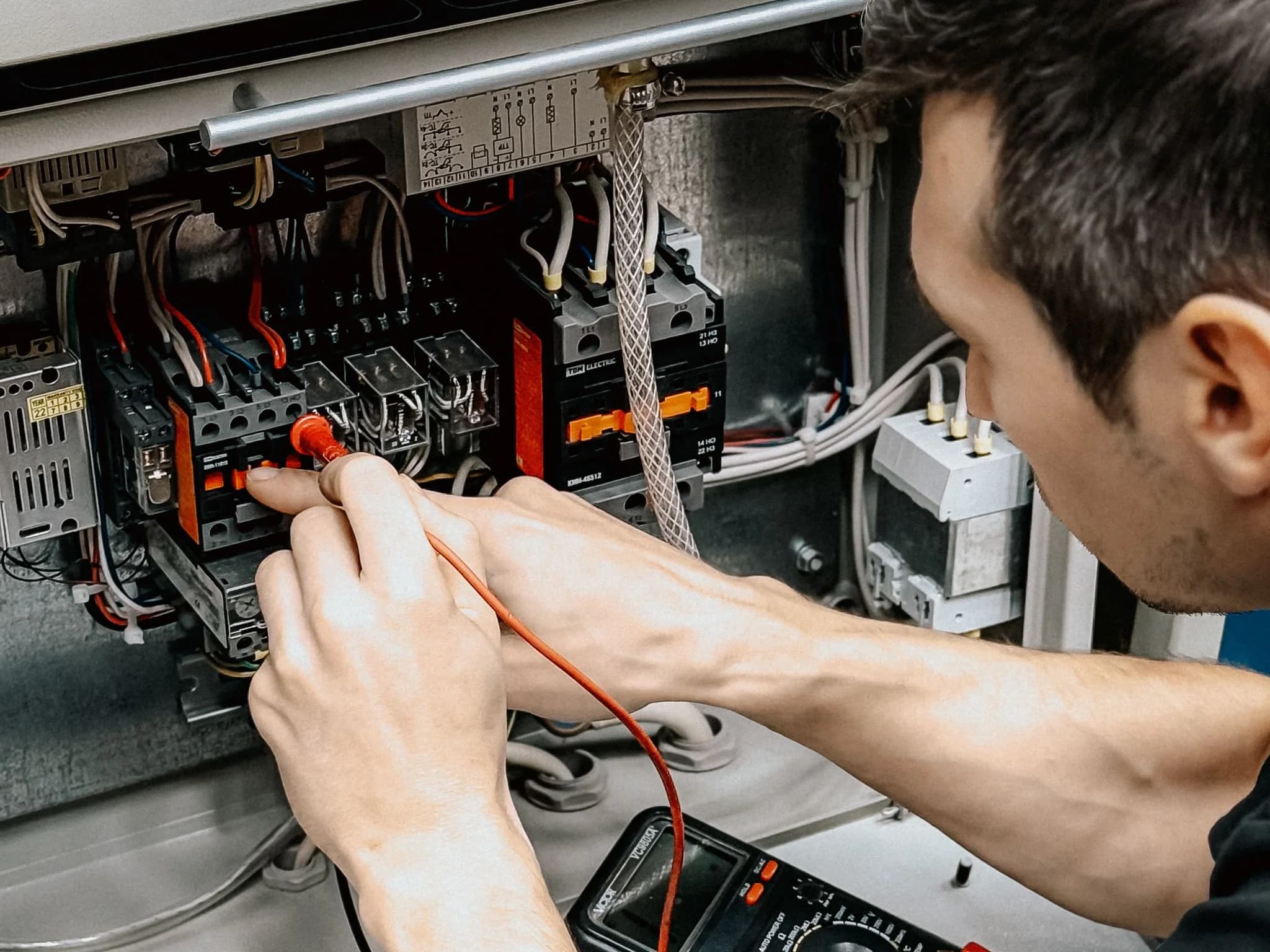 An electrician testing inside an open control panel with a probe, a multimeter on the bench below